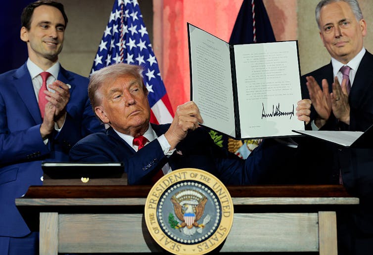 Man wearing suit holds up a padfolio featuring his signature as he's flanked by two men wearing suits who are clapping.