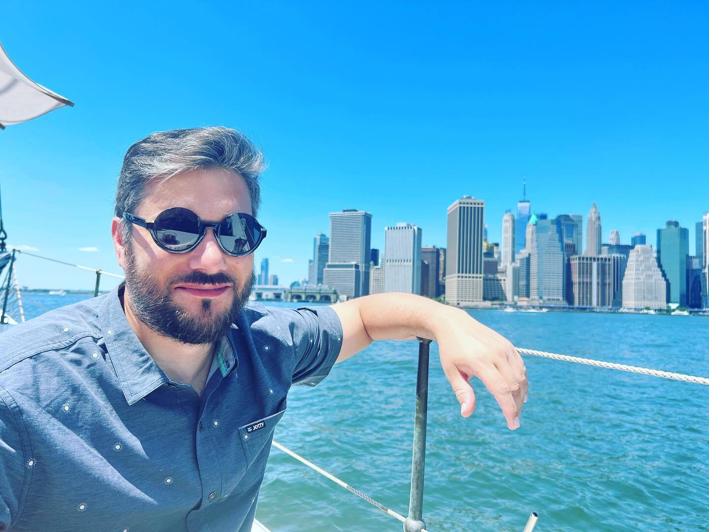 a man in sunglasses sits on a boat in front of the lower Manhattan skyline
