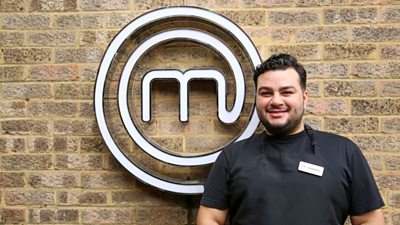 A portrait image of a contestant stood against an exposed brick wall, with the MasterChef circular white logo behind him.
