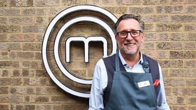 A portrait image of a contestant stood against an exposed brick wall, with the MasterChef circular white logo behind him.