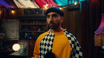 Image of Mawaan Rizwan looking at something off camera. Mawaan is wearing a yellow and black and white check jumper