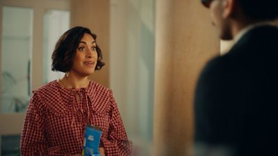 Emily Lloyd Saini, who plays Winnie, talking to someone off camera. She is wearing a red check blouse.