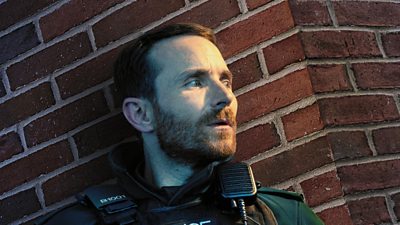 Martin McCann in character as Stevie Niell on set of Blue Lights stood up against a red brick wall waiting for something. He is in police uniform.