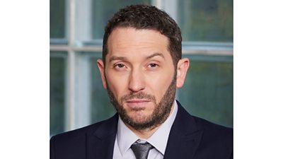 A head and shoulders shot of a man in a suit, looking to camera with an eyebrow raised