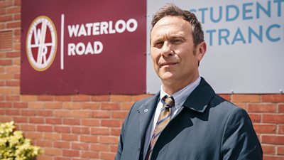 A man stands outside a school in the sunshine, wearing a dark coat over a shirt and tie.