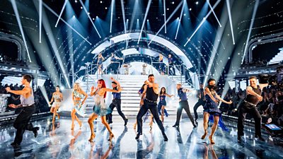 The Strictly professionl dancers move energetically across the dance floor in formation, lit by bright white lights