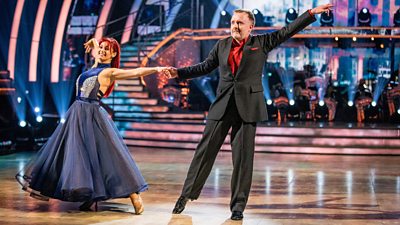Dianne Buswell and Chris McCausland dancing - they are linked by a single hand as they stretch apart mid routine in an elegant ballroom dance
