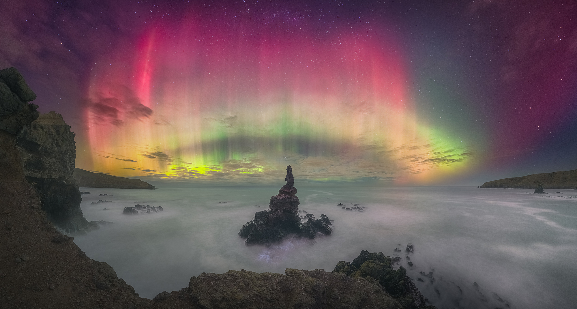 Bright red, green and yellow aurora are pictured swirling through the starry sky above a rocky bay leading out to the ocean. The surface of the water appears misty, and a spindly rocky formation is positioned in the middle of the image, appearing to reach up towards the aurora.