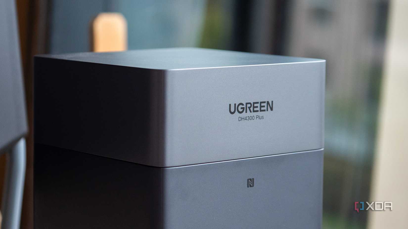 Dark gray UGREEN DH4300 Plus NAS, blurred background with furniture.