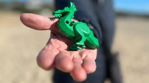 BBC A close-up of a hand holding a small green toy dragon. The background is blurred and the person is wearing dark clothing.
