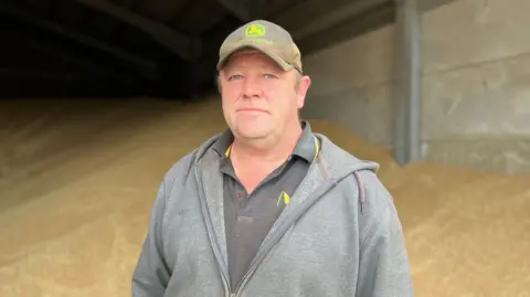 BBC Dan stands in front of a pile of grain, he is wearing a grey zip hoodie with a grey polo shirt with yellow trim. He is wearing a light grey baseball cap.