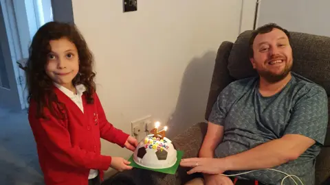 Natalie Morris A young girl in a red jumper presents a man sitting in a chair with a football birthday cake with candles in the shape of a 44. 