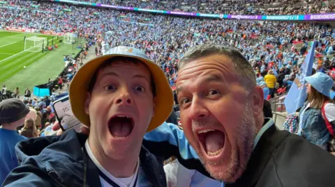 Andy Beaufoy and Richie Suthers Two men smiling toward the camera, behind them are football fans. They both have their mouths wide in an excited look.