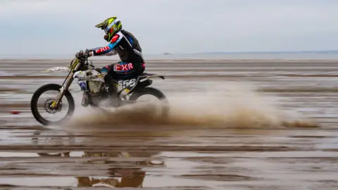 PA Media A person rides a white dirt bike with the number 556 along weston beach. It is muddy with water being thrown up by the bike. The rider wears a black red and blue kit and yellow and black helmet