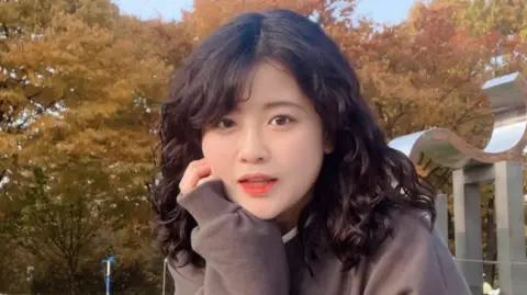 Instagram / Bae Se-hee Baek Se-hee resting her cheek on her hand as she looks at the camera. She has curly black hair and is wearing a brown sweater. In the background are trees with yellow leaves.