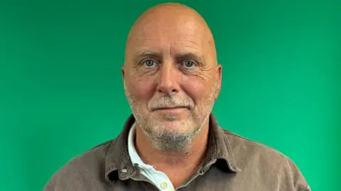 BBC Marco Catena has a short grey beard and is wearing a white polo neck shirt under a brown shirt.