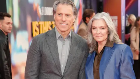 PA Media John and Melanie Bishop attending the screening of Is This Thing On? in London. John, with grey hair, is wearing a grey suit and lighter grey shirt. Melanie, with longer grey hair, is wearing a blue jacket and brown shirt. They are both smiling. 