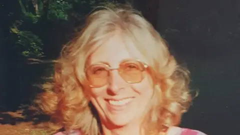 Handout A faded image of a woman with curly blonde shoulder-length hair and tinted sunglasses smiling. Bright sun is shining in her face, and the woodland behind is partially in shadow. She is wearing a checked red and white dress.