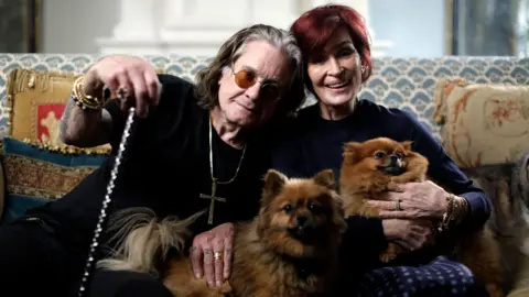 PA Media Ozzy Osbourne, who is pictured with long dark grey hair and wearing orange-tinted glasses and chunky gold jewellery, including a cross around his neck, sits on a blue and white patterned sofa with gold and blue cushions next to Sharon Osbourne, who has short bright red hair and is smiling. They are holding two small dogs.
