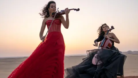 Sally Zarzour Two women outdoors in the desert, playing the violin and the cello. One woman is standing up, wearing a red dress, the other is sitting down and wearing a black dress.