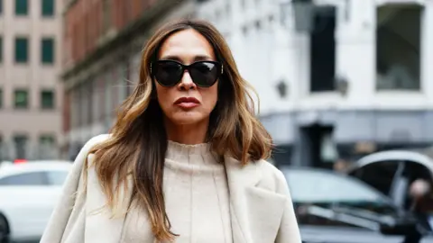 PA Media A woman with long brown hair is pictured wearing large black sunglasses and a white jumper and white coat. 