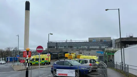 BBC Several ambulances and a couple of cars parked outside Derriford Hospital in Plymouth on a gloomy day. A large chimney is in the background.