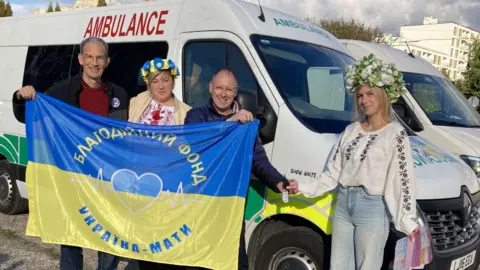 Towcester Tove Benefice Two white ambulances, in a car park, with four people standing by it, three are holding a flag. Two women have flowers on their head. 