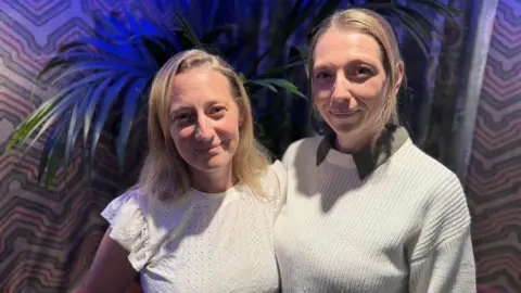 BBC Lauren and Katy Falla looking at the camera with slight smiles. Lauren, who is on the left hand side of the picture, has blonde shoulder length hair. She is wearing a white top with frilled sleeves. Katy, who is on the right hand side of the picture, is brown-blonde hair which is tied back. she has a white jumper on and you can see green collars from a shirt underneath.