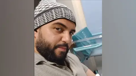 Facebook A selfie of a man who is sitting down in a hospital. He is looking directly at the camera. He is wearing a beige polo shirt and wearing a black and is wearing what appears to be a white knitted kufi hat.