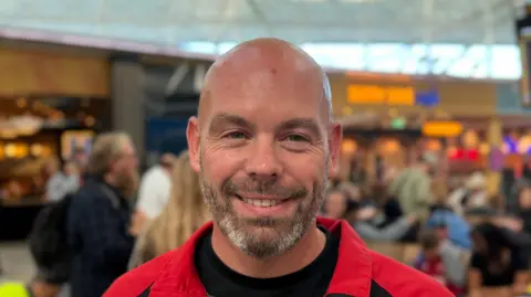 Elliot Deady/BBC Adam is standing inside Stansted Airport. There are people in the background as well as shops and restaurants.
