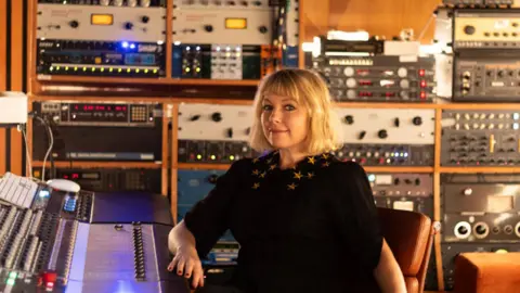 Bath Spa University Victoria Mason wearing a black dress with puffy sleeves that go down to her elbows, and gold stars on the collar. She is sitting in an orange leather chair with her elbow resting on a producer's desk. She appears to be in a recording studio, with lots of large technical equipment on wooden shelves filling the whole wall behind her, with buttons and dials and light-up switches. Victoria has light blonde hair which goes just down to her shoulders, with slightly darker routes and a short block fringe. She is wearing dark eyeliner and red nail varnish, and is smiling at the camera.