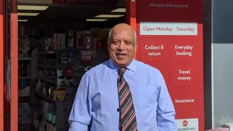 Post Office Bill Chandi, wearing a light blue shirt and striped tie, stands outside his shop, smiling. The door is open and to the right of it is red Post Office branding.