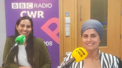 BBC Two women smile into the camera behind microphones. One on the left has long dark hair past her shoulders and a green jacket over a white top. The microphone in front of her is green. The woman on the right has a blue head scarf tied around her head and a white and black vertically striped jacket. The microphone in front of her is yellow.