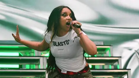 Getty Images In this photo, JADE is on stage at Glastonbury. She is singing holding a microphone in one hand and her other arm extended out. She is standing in front of stage risers, wearing a t-shirt that says "GLASTO".