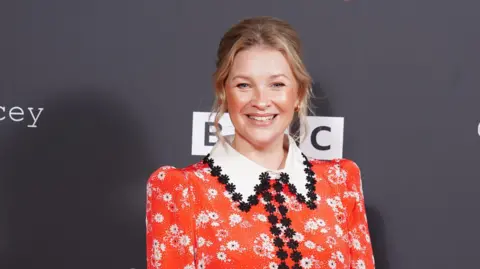 PA Media Joanna Page at launch event for Gavin and Stacey finale poses for photo in front of black background which says BBC behind her wears a red floral dress with large white collar and black floral embroidery around the collar and down the middle.