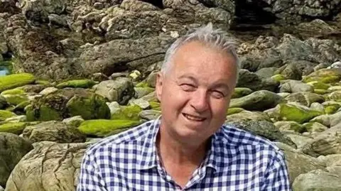 Grant Aldred Grant Aldred is sitting down with rocks behind him. He has short grey hair and is wearing a blue and white checked shirt with an open collar