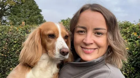 Paul Seager A smiling woman with shoulder length brown hair looks to camera. She is holding up a ginger coloured spaniel dog alongside her. She is stood in front of a green hedge.