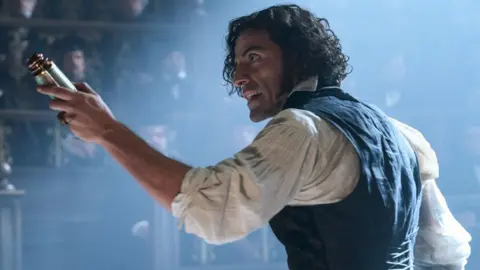 Netflix Oscar Isaac in white shirt and black waistcoat holds a piece of brass machinery in his left hand as he speaks to someone to his left. Men can be seen sitting in seats behind him. He looks passionate and has long black hair.