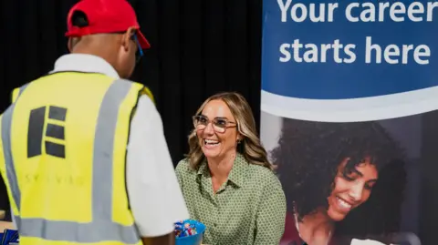 Darogan A woman in a green shirt and wearing glasses smiling at a man in a high vis vest while sitting at a desk