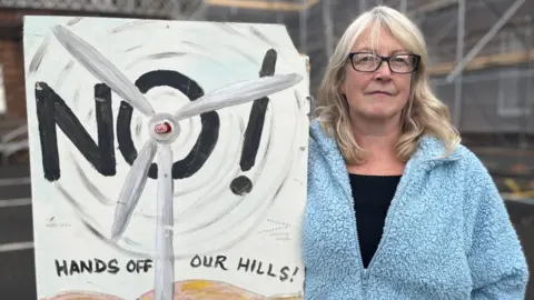 BBC A woman in a light blue fleece top and black T-shirt with long blonde hair and glasses holds up a "Hands Off Our Hills" placard