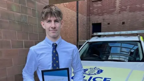 Police Scotland Dumfries and Galloway A male teenager wearing a blue shirt, black trousers, and brown shoes, stands in front of a police car. He is holding a certificate.