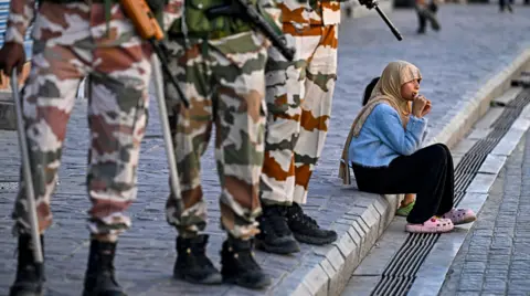 AFP Indo-Tibetan Border Police (ITBP) personnel stand guard near closed shops as a girl enjoys a chocolate along a pavement after curfew was partially relaxed for a few hours in Leh on September 27, 2025. 