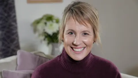 BBC Presenter Mari Grug smiling. She is wearing a purple jumper and her short, dyed blonde hair is tied back. She is sitting on a sofa, which has light-coloured purple cushions.