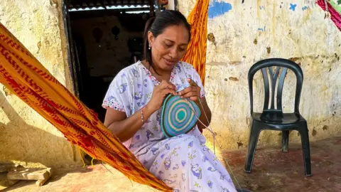 Yamile Vangrieken works on making a bag as she sits in an orange and red coloured hammock