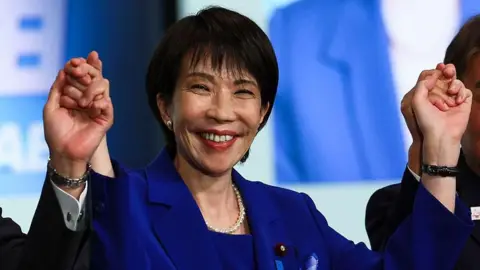 Getty Images Sanae Takaichi grins as she wears a blue dress with both her hands raised.