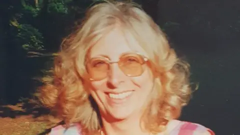 Handout A faded image of a woman with curly blonde shoulder-length hair and tinted sunglasses smiling. Bright sun is shining in her face, and the woodland behind is partially in shadow. She is wearing a checked red and white dress.