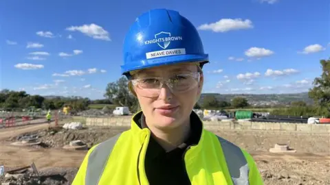 BBC Jack Little on a construction site, wearing a high vis jacket, a blue hard hat, and protective glasses. 