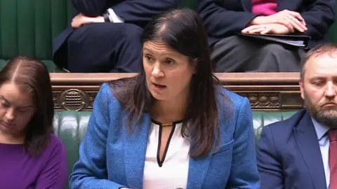 UK Parliament Lisa Nandy, with long dark hair and wearing a blue jacket, addresses the House of Comments flanked by colleagues on the green benches.