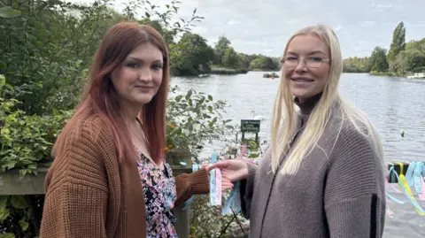BBC A woman with red hair and wearing a brown cardigan stands by a lake with another blonde woman, who is wearing a grey cardigan. Both women are holding a blue and pink ribbon in their hand. 