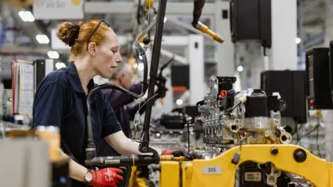 Jaguar Land Rover A female worker operates machinery at Jaguar Land Rover's factory in Wolverhampton. She has red hair which is tied back and has glasses perched on her head. She is wearing a dark top and red gloves.